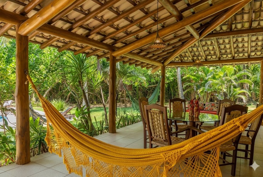 Varanda coberta com mesa de almoço e rede amarela, jardim ao fundo